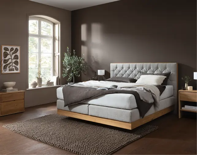 Schlafzimmer mit gepolstertem Bett, Nachttischen aus Massivholz und warmem Wandton. Hochwertige Schlafzimmermöbel mit natürlichem Design, Teppich und großem Fenster für gemütliches Wohnen und erholsamen Schlaf.